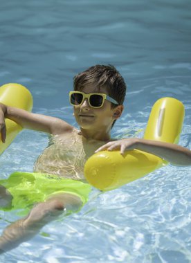 Güneş gözlüğü takan genç çocuk yaz gününün tadını çıkarıyor. Yüzme havuzunda sarı şişme üzerinde dinleniyor.
