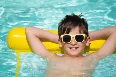 Kaygısız çocuk yazın tadını çıkarıyor, ferahlatıcı mavi yüzme havuzunda sarı şişme bir yatakta yüzüyor..