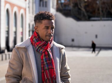 Kırmızı eşarp ve bej ceket giyen şık siyah adam güneşli bir kış gününde şehirde yürüyor.