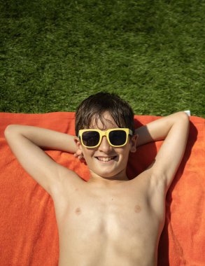 Güneş gözlüklü, dışarıda havluyla rahatlayan gülümseyen çocuk.