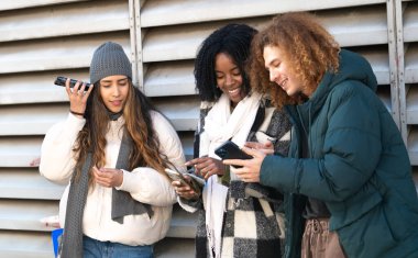 Üç genç öğrenci cep telefonlarını kullanıyor ve kış kıyafetleriyle dışarıda sosyalleşiyorlar.
