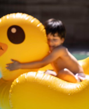 Küçük çocuk güneşli bir havuzda şişme ördeğe sarılıyor.