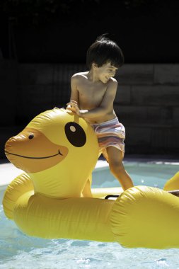 Küçük çocuk yüzme havuzunda dev bir şişme sarı ördeğe binerek eğleniyor..