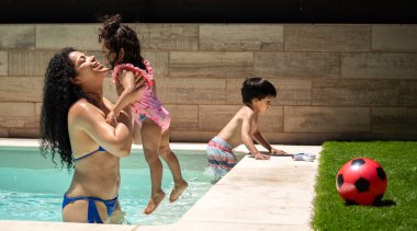 Annesi gülümseyen kızını arka bahçedeki yüzme havuzuna kaldırırken oğlu yakınlarda oynuyor.