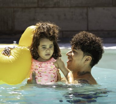 Babası kızını yüzme havuzunda tutarken o da şişme ördekle oynayıp güneşli bir günün tadını çıkarıyor..