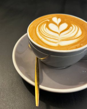 Gri seramik fincanda latte sanat kahvesi, üzerinde altın bir kaşık, koyu bir yüzey üzerinde dinleniyor.