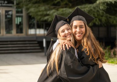 İki mutlu mezun öğrenci üniversitelerinin önünde birbirlerine sarılıyorlar.