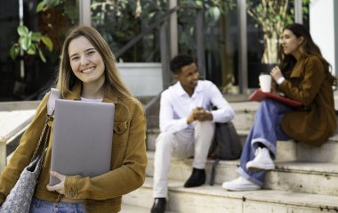 Dizüstü bilgisayarı olan mutlu bir üniversite öğrencisi, gülümsüyor, sınıf arkadaşları arka planda oturuyor.