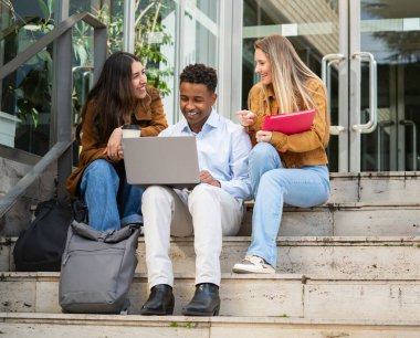 Bir grup öğrenci, bir dizüstü bilgisayar kullanarak bir üniversite binasının merdivenlerinde iş birliği yapıyor..