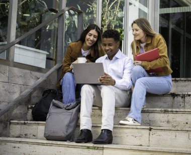Üniversite binasının dışındaki basamaklarda otururken dizüstü bilgisayar kullanan ve proje üzerinde işbirliği yapan mutlu öğrenciler