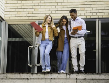 Üç mutlu çok ırklı öğrenci okul binasının önünde birlikte yürüyor, ellerinde kitap ve kahve tutuyorlardı.