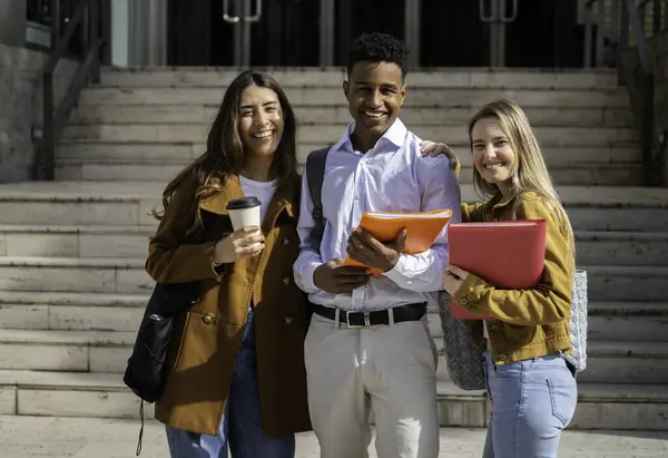 Üç neşeli çoklu etnik üniversite öğrencisi kampüs merdivenlerinde birlikte duruyor, ellerinde kitaplar ve kahveyle akademik hayatlarının tadını çıkarıyorlar.