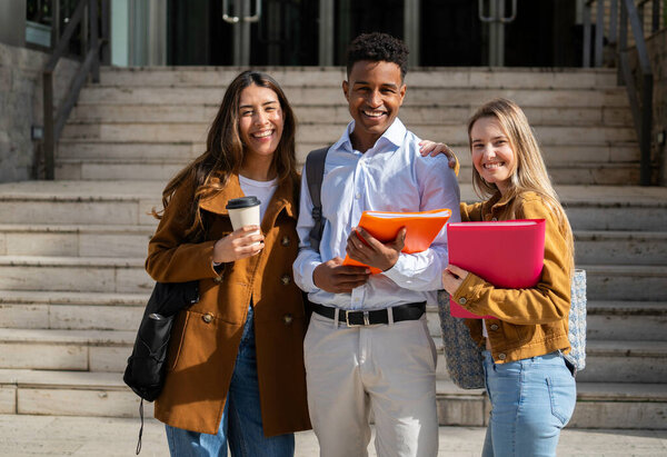 Три весёлых многонациональных студента колледжа стоят вместе на ступеньках кампуса, держа книги и кофе, наслаждаясь своей академической жизнью
