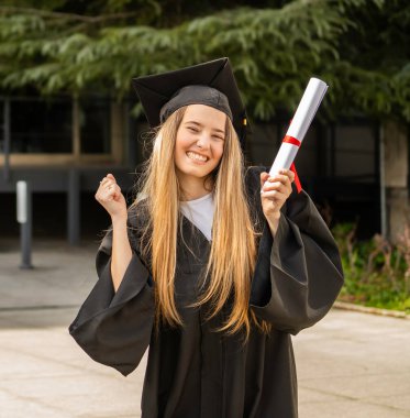 Heyecanlı öğrenci mezuniyeti kutluyor, diplomasını tutuyor ve mezuniyet elbisesi ve havan topu giyiyor.