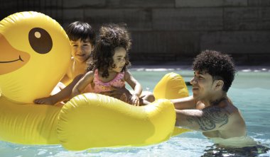 Baba ve çocuklar yüzme havuzunda eğleniyorlar. Kocaman, şişme sarı bir ördekle oynuyorlar.