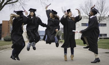 Bir grup neşeli, etnik çeşitliliğe sahip mezunlar önlük ve kep giyip diplomalarını alarak dışarı atlıyorlar.