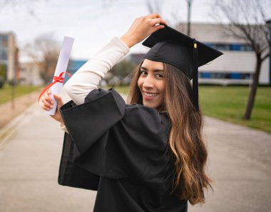 Mezuniyetini kutlayan, diplomasını tutan ve şapkasını gülümseyerek düzelten genç bir kadın.
