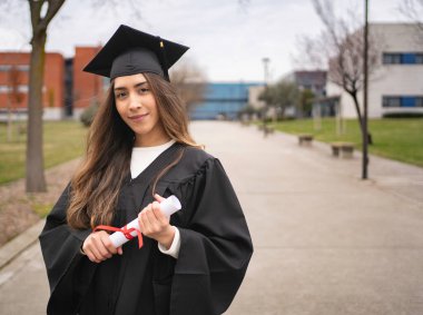 Üniversite kampüsünde yürürken gülümseyen ve diplomasını tutan gururlu bir yüksek lisans öğrencisi.