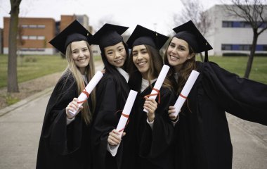 Dört gülümseyen bayan mezunlar üniversite kampüsünde diploma taşıyorlar.