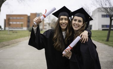 Mezuniyet elbisesi giymiş gülümseyen iki kız üniversite kampüsünde kucaklaşıyor ve diplomalarını tutuyorlar.