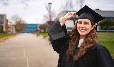 Akademik elbise giyen, elinde mezuniyet şapkası olan ve üniversite kampüsünde gülümseyen neşeli öğrenci portresi.