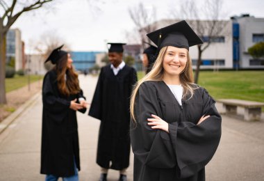 Üniversite kampüsünde arka planda sohbet ederken kollarını kavuşturup gülümseyen kendinden emin mezun.