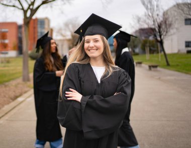 Kampüste kollarını kavuşturup gülümseyen genç lisansüstü öğrencisi ve arka planda diğer öğrenciler