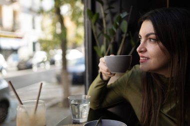 Genç bir kadın bir fincan kahveden zevk alıyor ve bir kafede pencereden dışarı bakıyor, ara veriyor ve şehir hayatını seyrediyor.