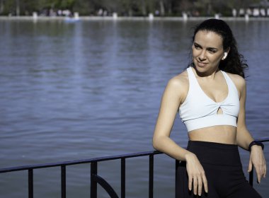 Kulaklık takan genç sporcu kadın, göl kenarındaki korkuluklara yaslanıyor, egzersiz yaptıktan sonra manzaranın tadını çıkarıyor..