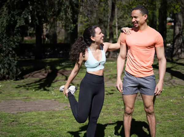 Gülümseyen sporcu kadın parkta onu destekleyen antrenörüyle bacak esnetiyor.