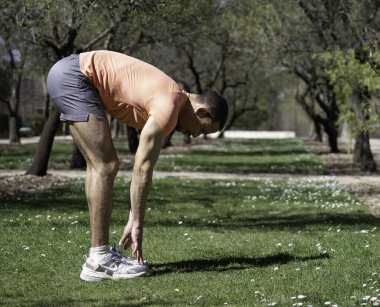 Spor giysili genç adam koşmadan önce ağaçlarla dolu bir parkta bacaklarını açıyor.