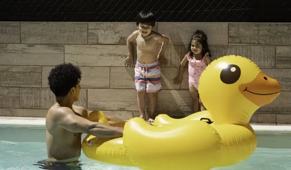 Baba, yaz boyunca oğlu ve kızıyla yüzme havuzunda dev, şişme sarı bir ördekle oynuyor..