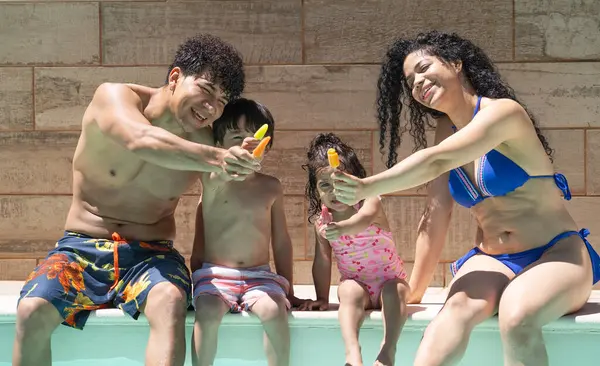 İki çocuklu gülümseyen aile yaz tatilinde yüzme havuzunda otururken renkli dondurma yiyorlar.