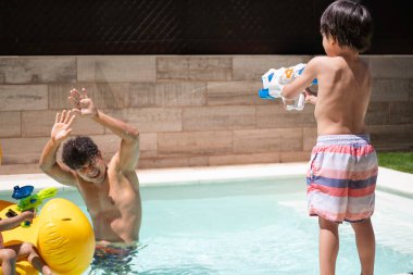 İspanyol çocuk güneşli arka bahçede su tabancasıyla eğleniyor ve babasını vuruyor.