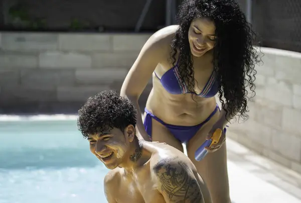 Gülümseyen Latin kadın güneşli bir yaz gününde erkek arkadaşına havuz kenarında güneş kremi sürüyor.