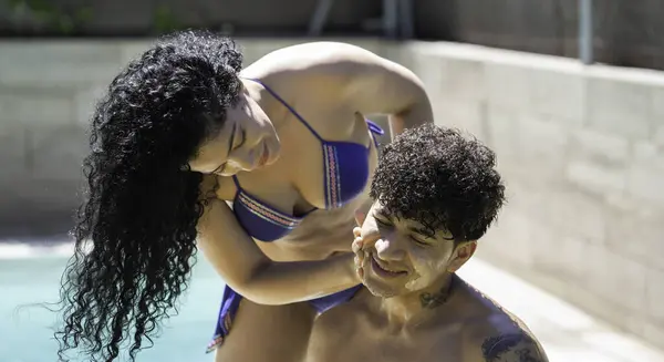 Gülümseyen genç Latin kadın güneşli bir günde genç bir adamın yüzüne dikkatlice güneş kremi sürüyor.