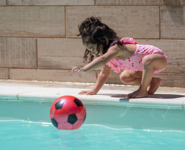 Havuzun kenarında çömelmiş, şişme futbol topunu suya iten genç Latin kız.