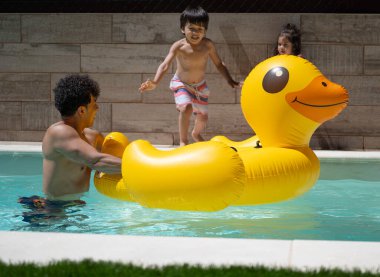 Baba, oğluyla ve kızıyla yüzme havuzunda kocaman, şişme sarı bir ördekle oynuyor.