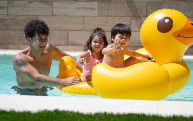 Latin baba, oğlu ve kızıyla eğleniyor. Yaz boyunca yüzme havuzunda dev, şişme sarı bir ördekle oynuyor.