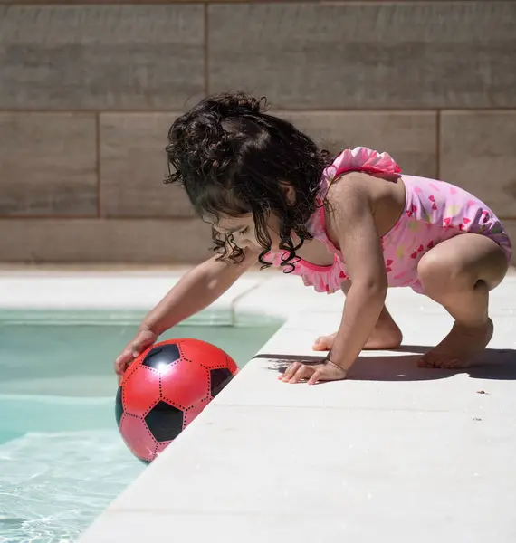 Profil görüntüsü pembe mayo giymiş Latin kız şişme futbol topunu güneşli yaz gününde arka bahçeye yüzme havuzuna koyuyor.