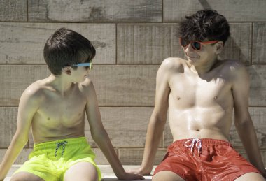 Neşeli genç çocuklar havuz kenarında güneşli bir günün tadını çıkarıyorlar, renkli mayo giyip birbirlerine bakıyorlar.