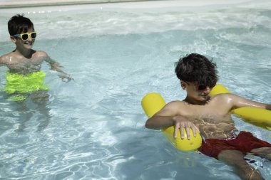 İki çocuk yaz tatilini yüzme havuzunda geçiriyorlar. Biri yüzüyor, diğeri ise sarı şişme havuzda yüzüyor.