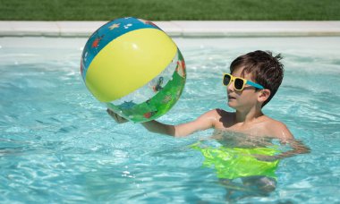 Çocuk güneş gözlüğü takıyor ve yaz tatilinde havuzda yüzerken şişme top tutuyor.