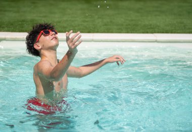 Kırmızı güneş gözlüğü takan genç bir çocuk yaz boyunca fotokopi odasıyla yüzme havuzunda su damlalarıyla oynuyor.