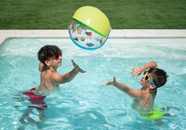 Güneş gözlüğü takan iki genç çocuk güneşli bir yaz gününde yüzme havuzunda renkli şişme bir topla oynuyorlar.