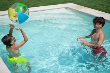 Güneş gözlüğü takan iki genç yaz tatilinin tadını çıkarıyorlar. Yüzme havuzunda fotokopi alanı olan renkli bir şişme topla oynuyorlar.