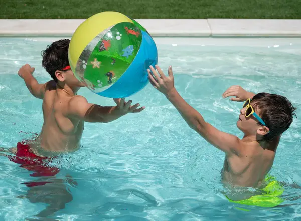 Güneş gözlüğü takan iki çocuk yaz boyunca yüzme havuzunda şişme topla oynuyorlar.