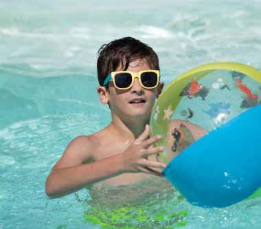 Güneşli bir yaz gününde yüzme havuzunda renkli şişme topla oynayan gülümseyen çocuğun portresi.