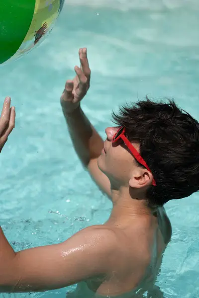 Güneş gözlüklü genç adamın yüzme havuzunda şişme plaj topuyla oynadığı yakın plan görüntüsü.