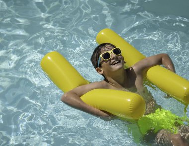Güneş gözlüğü takan neşeli genç çocuk yaz tatilinin tadını çıkarıyor, yüzme havuzunda sarı şişme üzerinde dinleniyor.